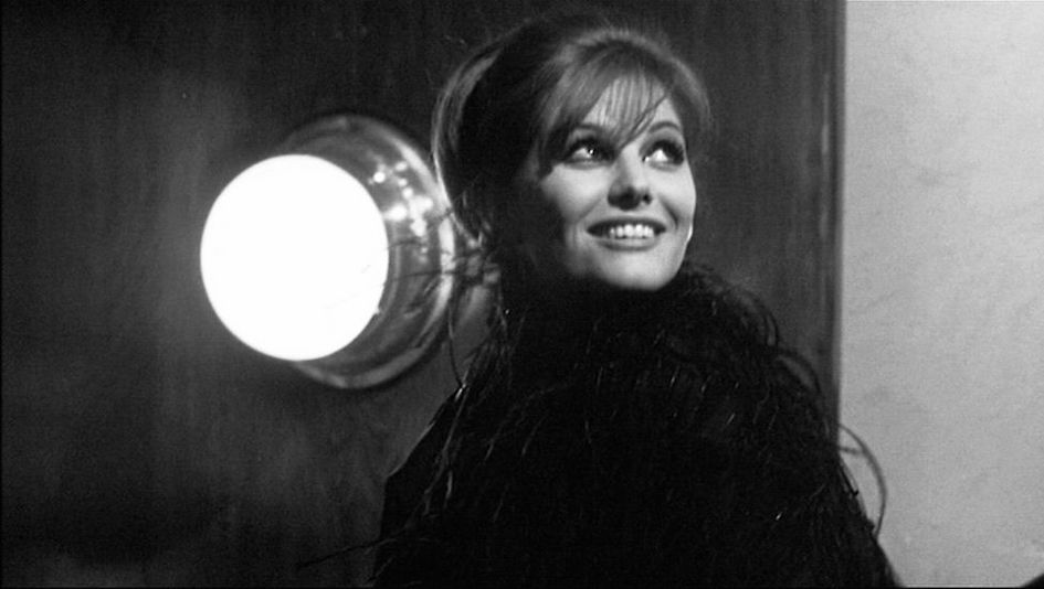 Film still from OTTO E MEZZO: A smiling young woman looks up at the sky, a bright lamp shines on the wall next to her.