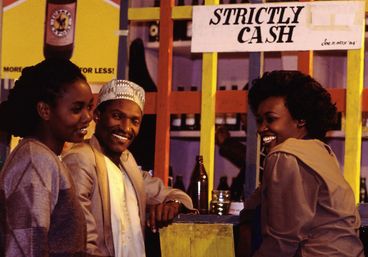 Still from the film „Der Kampf um den heiligen Baum" by Wanjiru Kinyanjui. Two women and a man standing at a bar laughing.