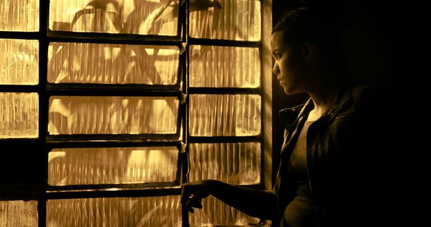 A young woman stands at the side of a window made of fluted glass and looks out. The image is bathed in yellowish light. 