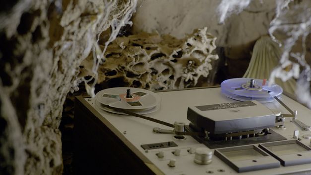 Film still from "Resonance Spiral" by Filipa César and Marinho de Pina. It shows a close-up of a tape recorder with a tape. 