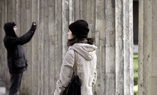 Still from the film „Onun Haricinde, yiyim“ by Eren Aksu. A row of pillars can be seen. In the front is a woman wearing a beanie and looking around. In the background is a person taking a photo.