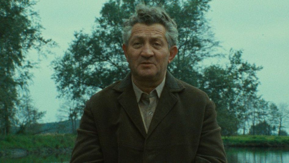 Film still from SHOAH: An elderly man in conversation, water and trees can be seen behind him.