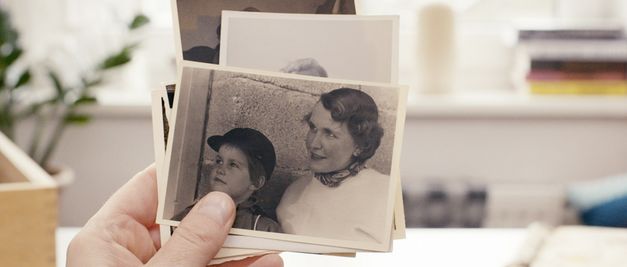 Film still from "Reproduktion" by Katharina Pethke. It shows a close-up of a hand holding photos. The front photo is a black and white picture of a child and a woman. 