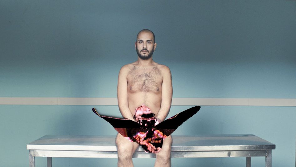 Filmstill aus EMBODIED CHORUS: Ein junger Mann sitzt nackt auf einer Bank vor einer türkis gestrichenen Wand. 