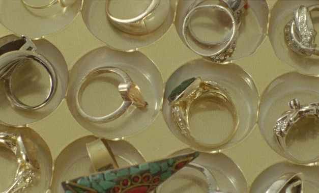 Still from the film "Mis Dos Voces" by Lina Rodriguez. We see a close-up of rings inside individual jewellery dishes, against a yellow background. 