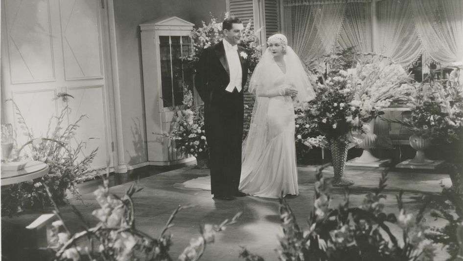Film still from DESIGN FOR LIVING: A wedding couple stands in a room full of flowers.
