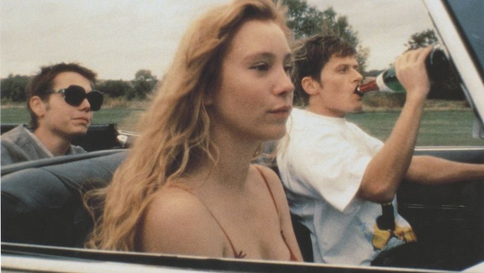 Film still from VERGISS AMERIKA: Three young people are sitting in a car, the driver is drinking champagne from a bottle.
