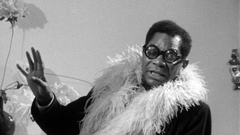 Film still from PORTRAIT OF JASON: A man with glasses and a feather boa in conversation.