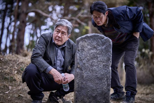 Film still from "Pa-myo" by Jang Jae-hyun. It shows two men standing by a gravestone in the middle of nature. The one on the left is looking closely at the stone, while the one on the right is looking past it. 