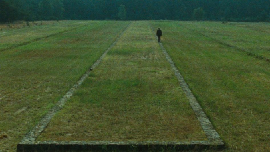 Filmstill aus SHOAH: Zu sehen ist eine Landschaft mit Wiese, im Hintergrund Wald. Auf der Wiese sind die Überreste einer Bebauung zu erkennen, eine Person geht daran entlang.
