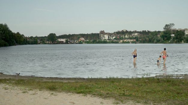 Film still from "Intercepted" by Oksana Karpovych. It shows a lake with four people in it. There are trees around the lake. Buildings can be seen in the distance. 