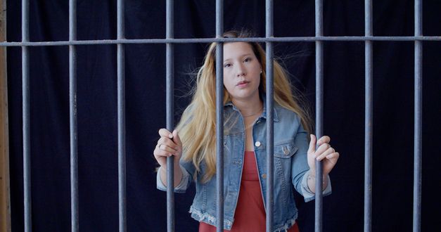 Film still from "Reas" by Lola Arias. It shows a woman in a denim jacket behind bars. 