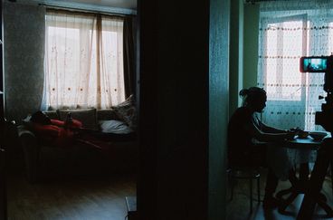 In the left of half the image a man wearing a red fur and leather jacket lounges on a couch, looking at his cell phone. On the right, in the adjacent room a man in silhouette sits at a kitchen table with his phone. To the far right we see a camera, with the second man framed in its composition.