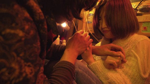 Film still from "Republic" by Jin Jiang. It shows a close-up of two people sitting close together and holding hands. 