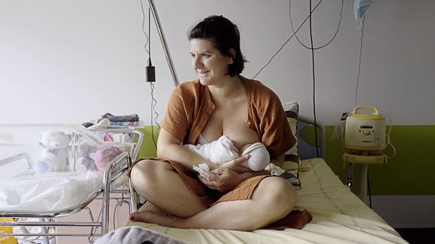Filmstill aus „Notre corps“ von Claire Simon. Das Filmstill zeigt eine lächelnde Frau, die auf einem Krankenhausbett sitzt und einem Baby die Brust gibt.
