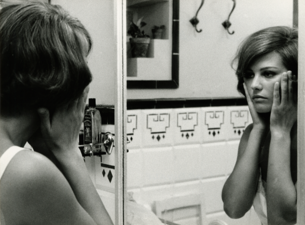 Film still from GLI INDIFFERENTI: A woman looks at herself in the bathroom mirror and holds her hands to her cheeks.