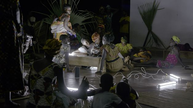 Film still from "Resonance Spiral" by Filipa César and Marinho de Pina. It shows people of different ages sitting outside at night. The scene is illuminated by white lamps. 