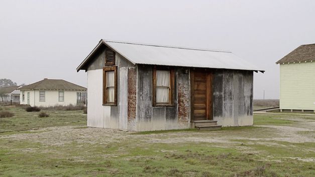 Filmstill from the movie "Allensworth" by James Benning. A wooden house in front of a grey sky. In the background some more houses.