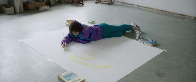 Film still from "Mit einem Tiger schlafen" by Anja Salomonowitz. It shows a person in a windbreaker lying on the floor on a large sheet of paper and painting. 