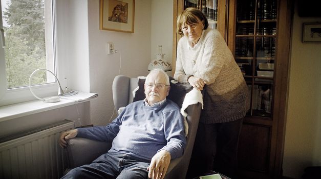 Still from the film "Gehen und Bleiben" by Volker Koepp. An elderly man in blue clothing, with grey hair and a grey beard, is sitting in an armchair. A woman with brown hair and a grey-brown cardigan is standing behinf him and leaning against the armchair. Both of them are looking at the camera. There is a dark wooden bookcase behind them.