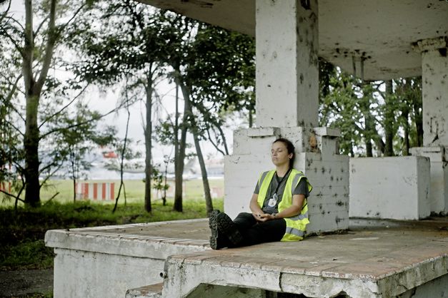 Still from the film "O Estranho" by Flora Dias and Juruna Mallon. A woman in a yellow vest sits on a stone platform with their eyes closed, leaning against a pillar. In the background, there are blurry trees and an airplane standing on an airport runway.