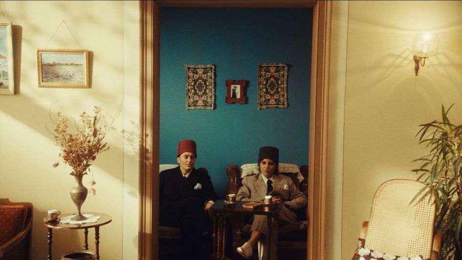 Film still from NEO NAHDA: Two women dressed as men sit between a door frame and look directly into the camera.