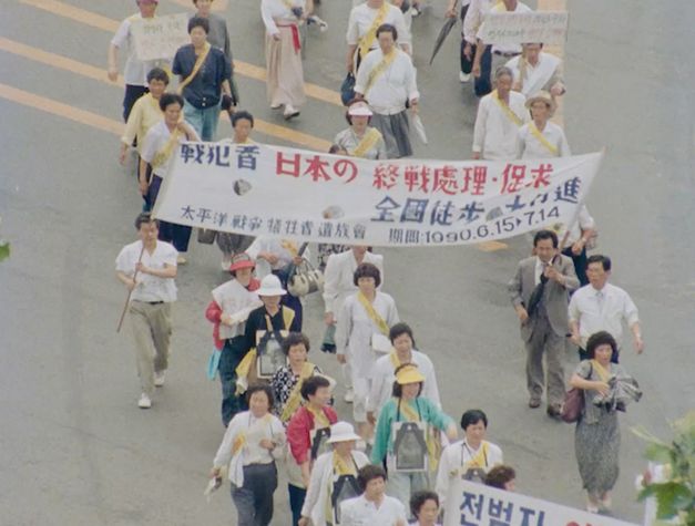 Film still from "Doesarananeun moksori / Yomigaeru Koe" by Park Soo-nam and Park Maeui. It shows a group of people walking together across the street in one direction, holding placards and banners.