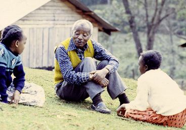 Still from the film „Der Kampf um den heiligen Baum" by Wanjiru Kinyanjui. Two children and a man sitting in a meadow and talking. In the background is a wooden cabin.