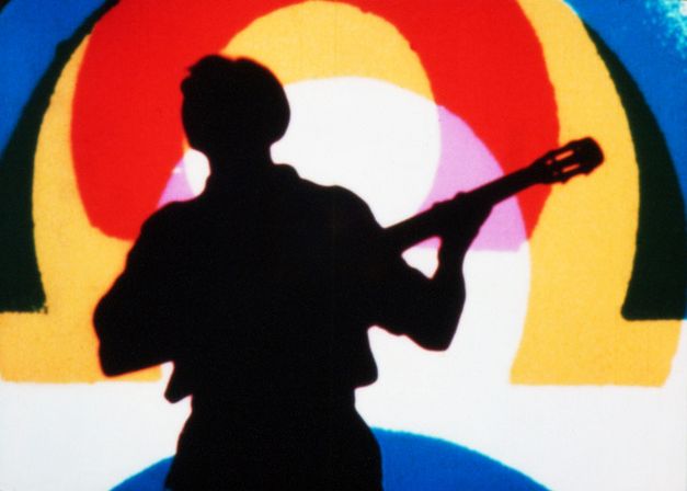 Film still from RAINBOW DANCE: In the foreground the silhouette of a person playing a guitar, behind it an implied rainbow in yellow, pink, red, black and blue.