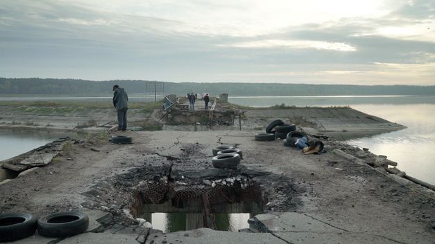 Film still from "Intercepted" by Oksana Karpovych.It shows a broken car bridge on a lake. A person is fishing at the edge of the bridge. Other people are walking in the background.