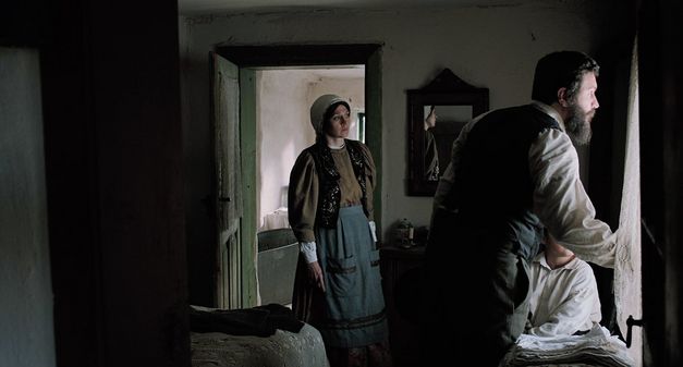Film still from "Săptămâna Mare" by Andrei Cohn. It shows a dark room with a man and a child at the window and a woman on the left in the doorway. They are all looking out of the window. 
