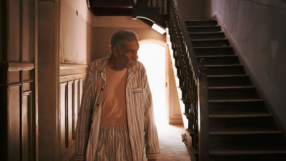 Film still from 19B: An elderly man stands in his pyjamas in a stairwell.