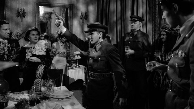 Film still from "Marijas klusums" by Dāvis Sīmanis. It shows a dinner party. In the centre of the picture is a man in uniform holding a gun in the air. 