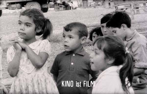 Film still from POR PRIMERA VEZ: A group of children are looking at the camera. Below is a lettering with the words cinema is film.
