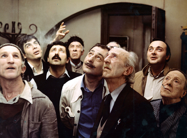 Filmstill aus BLAUE BERGE ODER EINE UNWAHRSCHEINLICHE GESCHICHTE: Eine Gruppe von Menschen steht in einem Raum und blickt nach oben.