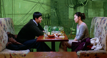 Filmstill aus TALE OF CINEMA: In einem Lokal sitzen sich ein Mann und eine Frau an einem Tisch gegenüber, der voller Gläser ist.