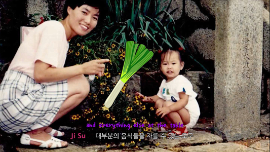 Filmstill aus KOREAN KAROTTENKUCHEN: Eine Frau und ein Kleinkind kauern zwischen Blumen auf dem Boden, zwischen ihnen ist ein Lauch montiert. Am unteren Bildrand sind Untertitel in Englisch und Koreanisch.