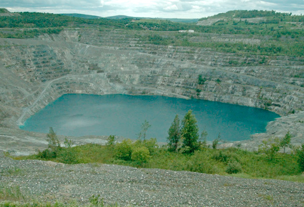 Still from “Asbestos“ by Graeme Arnfield and Sasha Litvintseva. A grainy image of a former asbestos mine. Surrounded by trees and rock, a huge whole filled with water has become a lake.