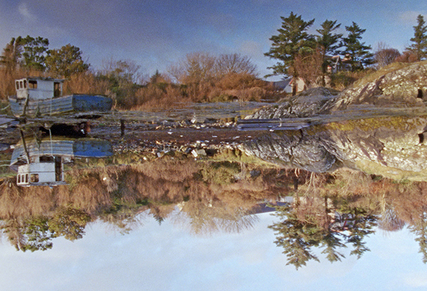 Filmstill aus „The Two Sights“ von Joshua Bonnetta. Bäume im Herbst, Felsen und ein gestrandeter Holzkahn, vom Wasser aus fotografiert. Im Wasser stillen spiegelt sich das Bild und die Landschaft taucht noch einmal über Kopf auf.