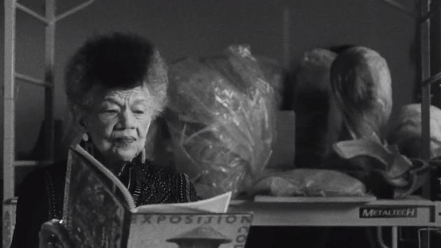 Film still from Simone Leigh and Madeleine Hunt-Ehrlich’s „Conspiracy“. On the left the torso of a person reading a magazine. In the background a shelf with objects wrapped in plastic on it.
