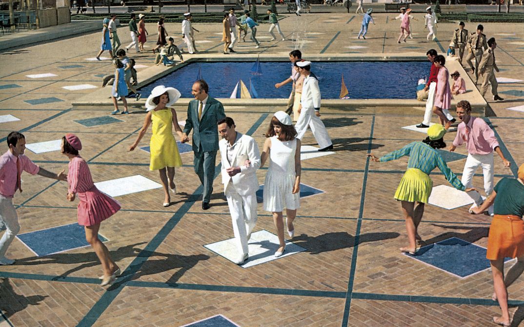 Filmstill aus LES DEMOISELLES DE ROCHEFORT: Viele lachende Menschen tanzen und laufen über einen Platz. Alle tragen auffallend bunte oder weiße Kleidung. In der Mitte ist ein Wasserbecken mit Miniatursegelbooten.
