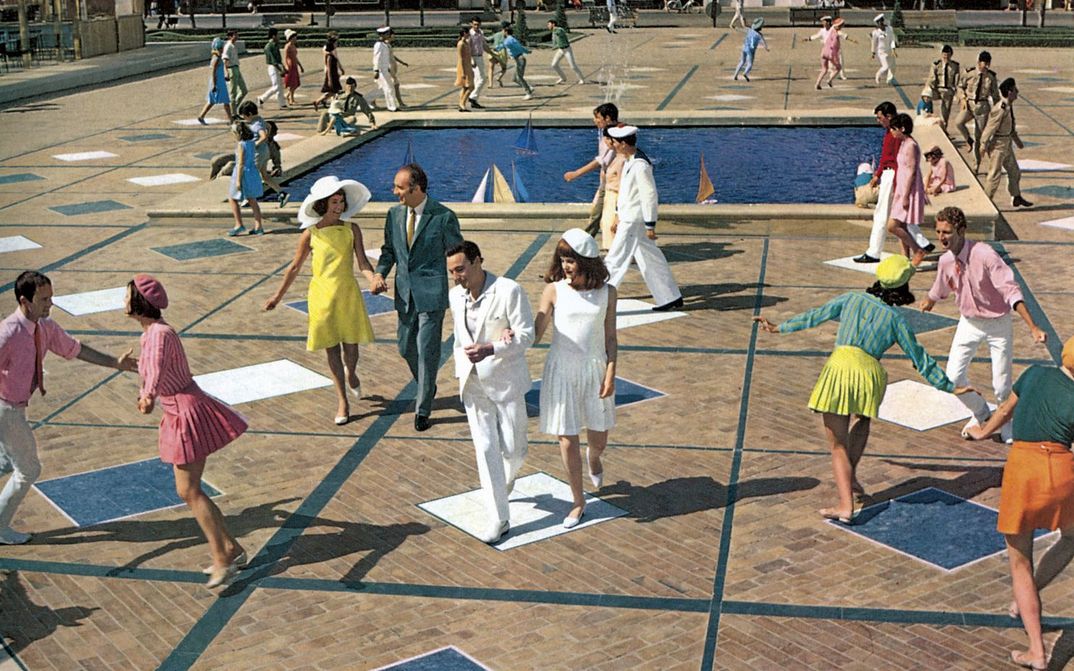 Filmstill aus LES DEMOISELLES DE ROCHEFORT: Viele lachende Menschen tanzen und laufen über einen Platz. Alle tragen auffallend bunte oder weiße Kleidung. In der Mitte ist ein Wasserbecken mit Miniatursegelbooten.