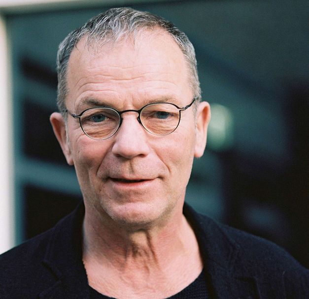 Portrait von Thomas Heise: Ein Mann mit kurzen grauen Haaren, einer Brille mit schmalem Rand. Er lächelt.