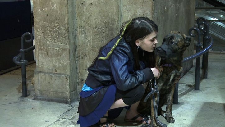 Film still from GEOGRAPHIE HUMAINE: A young woman crouches next to a dog on a street corner.