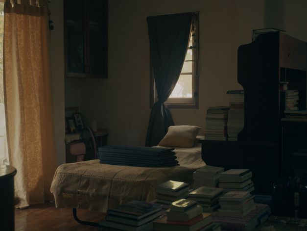 . Ein aufgeräumtes, schwach beleuchtetes Schlafzimmer. Das Bett ist gemacht, die Bücher sind ordentlich auf dem Boden und auf der Kommode gestapelt, und die Sonne lugt durch den dunklen Vorhang hervor.