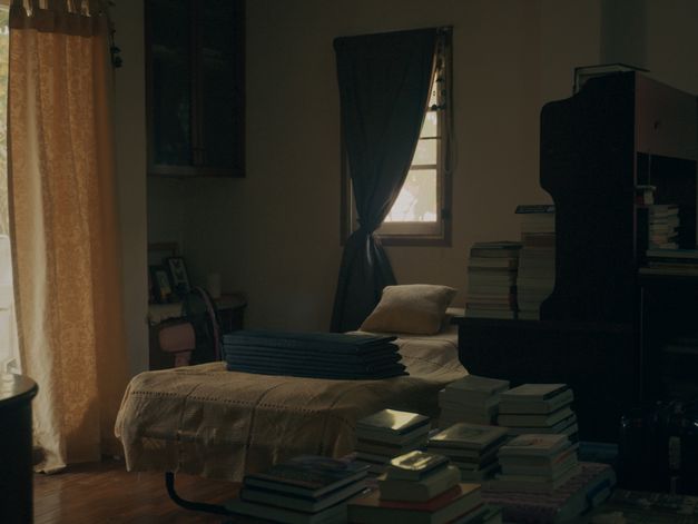 Film still from Chanasorn Chaikitiporn’s film “Here We Are”. An organised, low-lit bedroom. The bed is made, books are stacked neatly on the floor and on the dresser, and the sun peeps through the dark curtain. 