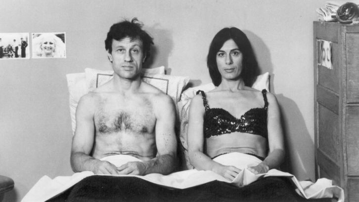 Filmstill aus KRISTINA TALKING PICTURES: Ein Mann und eine Frau sitzen nebeneinaner im Bett und schauen frontal in die Kamera.