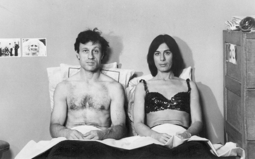 Filmstill aus KRISTINA TALKING PICTURES: Ein Mann und eine Frau sitzen nebeneinaner im Bett und schauen frontal in die Kamera.