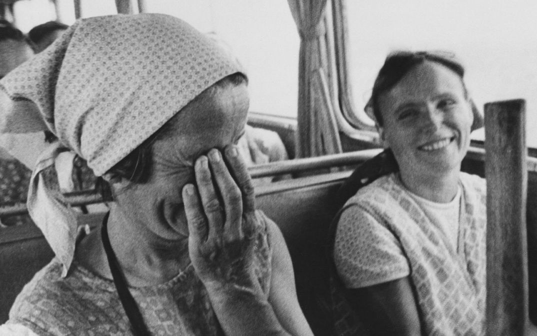 Film still from ON THE FIELD OF GOD IN 1972-73: Two women in peasant clothing are sitting in a bus. Both are laughing, one is holding her hand in front of her face.