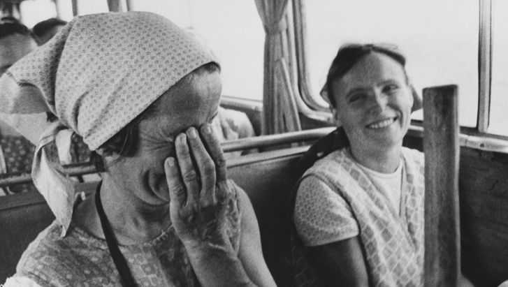 Film still from ON THE FIELD OF GOD IN 1972-73: Two women in peasant clothing are sitting in a bus. Both are laughing, one is holding her hand in front of her face.